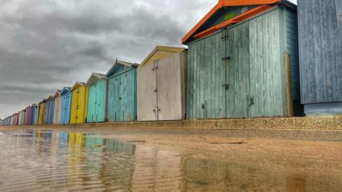 A row of coloured beach huts with grey cloudy skies behind and puddles in the sand in front