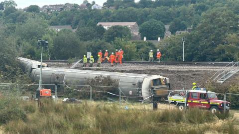 Three derailed train carriages lie at the bottom of a railway embankment near Lewes. Emergency vehicles, fencing and workers in high-visibility clothing can be seen around the site, while crews work on the railway line above.