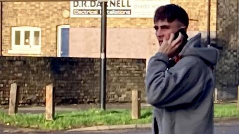 A dark-haired man in a grey hooded top holding a mobile to his left ear with his left hand. A building, a wall and a strip of grass are in the background