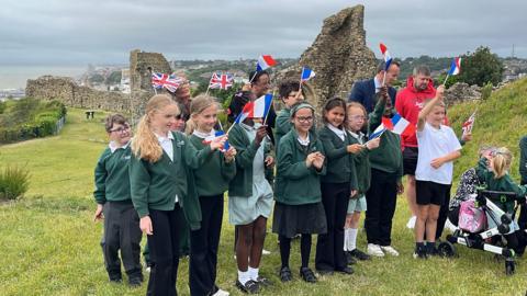 Primary school age children in green uniform waving Union Jacks and French flags on a cliff top with an ancient monument behind them