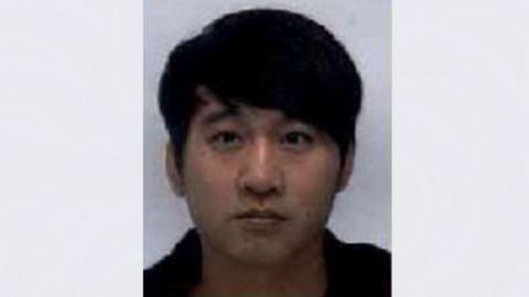 A grainy police mugshot of Zhaoqian Yu wearing a black hoodie and standing against a grey background. He has brown eyes and black hair styled in a thick sweeping fringe.