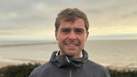 A man - Sam King - smiling straight at the camera. He is wearing a black running jacket and is standing in front of a beach in Walton-on-the-Naze.