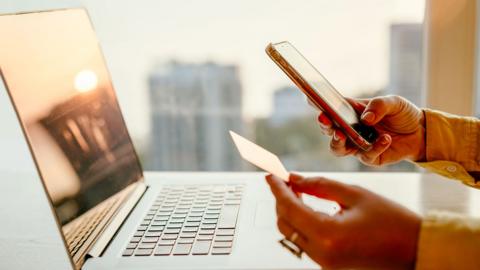 A woman in yellow blouse sitting at the table in front of a laptop and making online payment with a credit card. 