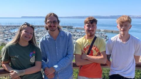 Four young people stood in a line. Left to right is Daisy Lloyd wearing a green top and black shorts, an unknown person wearing a blue top and grey shorts and sunglasses, Jarvis Rohr wearing a yellow and orange top and grey shorts and Lewis Jackson wearing a white top and black trousers. They are all smiling at the camera. Behind them is a harbour filled with boats and a ferris wheel and a bright blue sea and light blue sky.