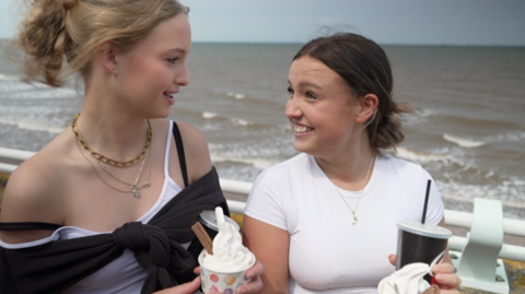 Two teenage girls turn to smile at one another. They are both holding Mr Whippy-style ice creams with chocolate flakes, in tubs. The sea is not far behind them. 