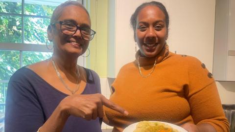 Sarah in a blue top pointing at a plate of doubles. She stands smiling next to her daughter Christina, who wears an orange roll-neck jumper. 