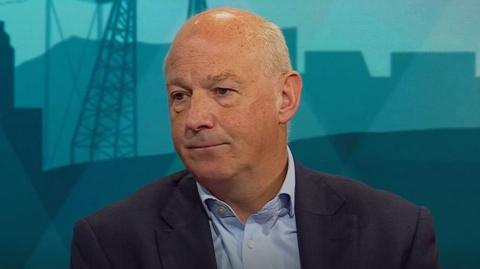 A man in a blue shirt and navy jacket sits on a sofa in a TV studio