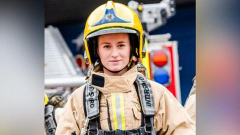 A female firefighter who is wearing all her protective clothing. She is wearing a yellow helmet and is smiling. 