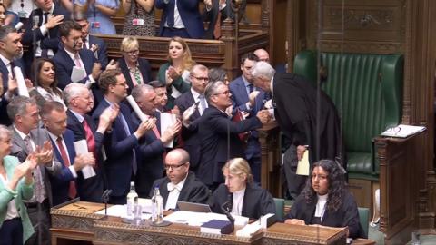 Keir Starmer shaking Lindsay Hoyle's hand surrounded by MPs applauding in the House of Commons.