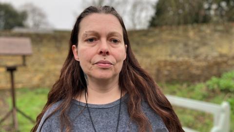 Jo, a woman, with long brown hair, wearing a grey jumper, looking into the camera. She is standing in a garden with grass in the background.