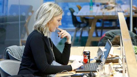 Woman with blonde hair sitting at a desk in front of a laptop in a well- lit office. She is wearing a black top and a pair of white in-ear headphones in her ears. She is holding her left index finger in her mouth while using the trackpad on the laptop keyboard.