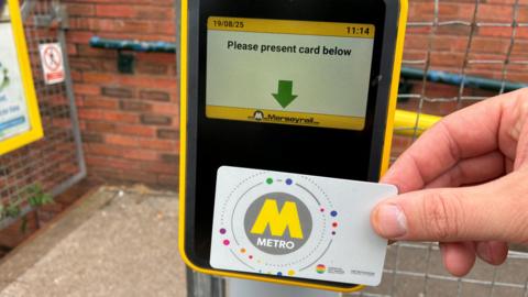 A close up of a hand holding a metrocard against a black scanner underneath a digital screen which says Please present card below.