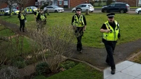 A still from doorbell camera footage showing six police officers walking up a path, roughly in a line, with a grassy area to their left. They are in black uniforms with yellow high-vis vests. The man at the front is pointing towards the camera or property.