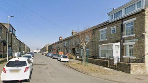 A quiet residential street lined with terraced houses on both sides. The houses are primarily built from light-coloured brick and have a traditional design, featuring rectangular windows and some with white-painted frames. Several cars are parked along the left side of the street, including a white car in the foreground and others further down the road.