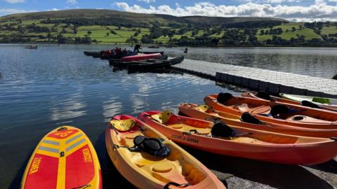 Caiacau yn arnofio ar Lyn Tegid tra bod plant i'w gweld yn rhywfo ar gwch arall yn y gorwel. 