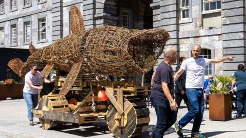 The wicker shark is being transported by a wooden trailer with wooden wheels. It is being guided at the front by two men and a woman is steering from the tail end area. They are pushing the model past old brick buildings. The model shark has its large mouth open as if it is feeding.