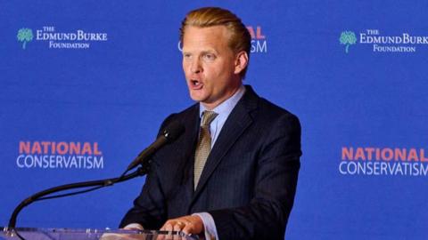 James Orr, a man with swept back blonde hair in a dark suit and sandy tie, speaks at a podium at a National Conservatism conference in Washington DC