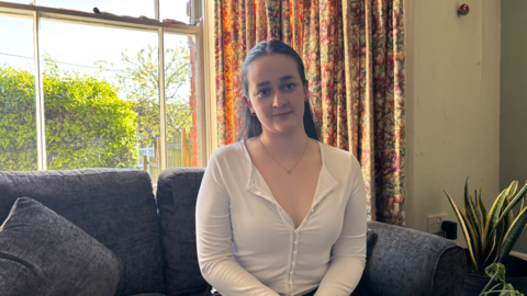 A portrait photo of a young woman in a white shirt with black hair - she has a slightly concerned look on her face. She's sitting on a sofa with a house plant to the right of the image. Behind her is a floral curtain hanging next to a window. Outside the window you can see a green hedge and a tree.