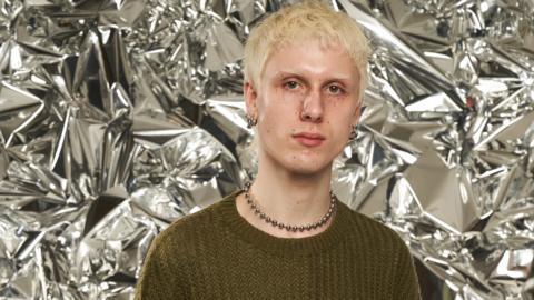 Jake Moran looks at the camera. He has short, bleach blonde hair. He is wearing a knitted green jumper and metallic necklace and silver hoop earrings. 