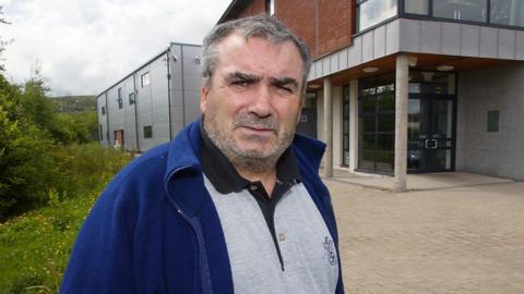 Freddie Scappaticci pictured in west Belfast in 2003 before he went into hiding in England. He is wearing a grey polo shirt under a blue fleece jacket.
He is looking into camera and has greying short hair and grey stubble. He is standing in front of a red brick building with panels of grey cladding.