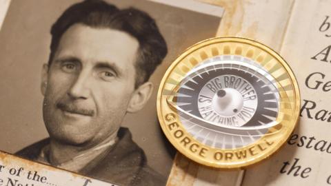 A handout photo issued by the Royal Mint of their new commemorative £2 coin to celebrate the life and legacy of George Orwell. It sits on top of a paper with Orwell's picture on and some writing. The coin's design - eye encircles the lens with inscription ‘BIG BROTHER IS WATCHING YOU’ and the name of the author.