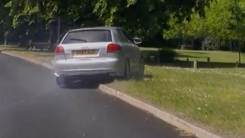 A silver Audi goes up on to a curb into a grassy park area.