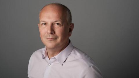 A professional-looking headshot of Simon Hewitt, a bald man wearing a light pink shirt. He's standing against a grey backdrop and looking straight at the camera.