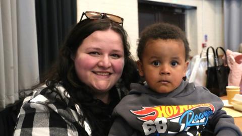 A woman in her mid-20s with shoulder-length dark hair and sunglasses on top of her head is smiling at the camera. She is wearing a white and black checked top. On her lap is her toddler son who is wearing a grey Hot Wheels Racing hoodie.