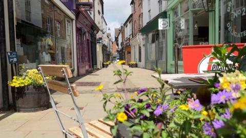 An empty street with shops either side of the path. In the foreground are plants and a table and chairs. Between the shops are beige slabs of paving.