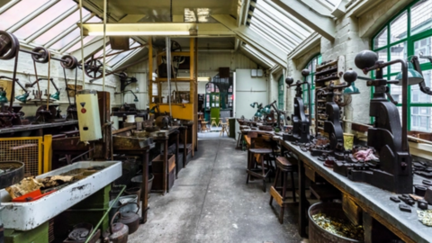 A jewellery workshop with old metal equipment on work benches, wooden stalls underneath and windows surrounding with green window panes