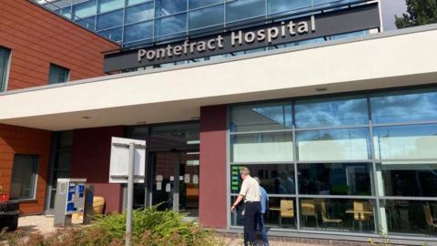 Two people walk into the entrance of a large modern-looking building. A sign out the front reads 'Pontefract Hospital'. 