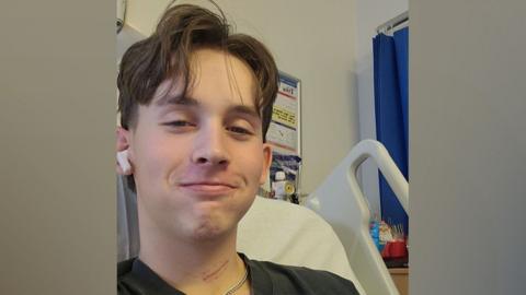 A picture of Ryan in hospital. He is taking a selfie style of photo and is smiling at the camera. He is lying in a hospital bed with headphones in his ears. He has brown, floppy hair that covers his forehead and he is wearing a black t-shirt.