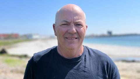 Donald Gillies stands with his back to a sandy coast. He is bald and wearing a navy blue t-shirt