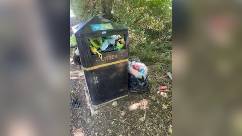 Overflowing bin. Bags of rubbish can be seen inside and on the floor next to the bin. 