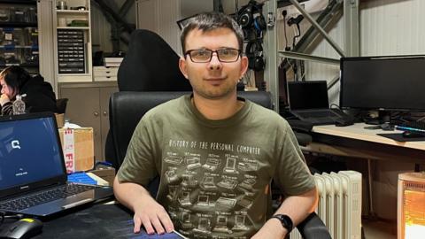 Cameron Clarke sits at a desk surrounded by computers. He has short brown hair and glasses and is wearing a green t-shirt covered in pictures of computers.