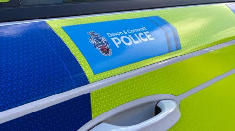 The side of a yellow and blue police vehicle. Above the door handle is a blue section with the words 'Devon and Cornwall Police' with a badge to its left. 