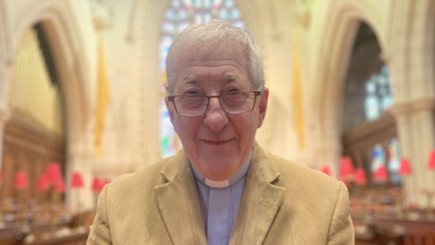 Shows a clergyman in a light-coloured jacket with a minister's blue garb underneath