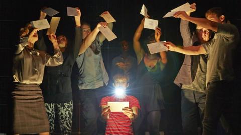 GCSE English literature image, The Curious Incident of the Dog in the Night-Time: Christopher holding a torch and reading a letter surrounded by the chorus holding letters