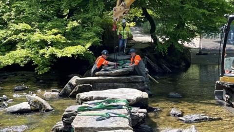 Workers in high-vis vests and hard hats use a digger to move huge slabs of stone crossing a shallow ford. Trees line the river bank. 