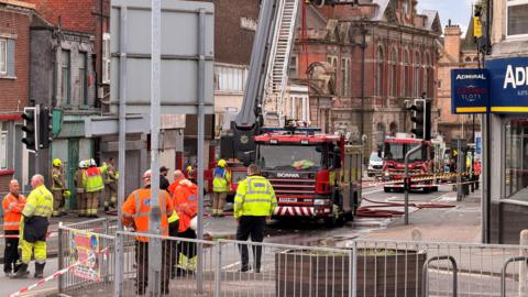 Emergency service personnel, wearing yellow or orange hi-vis jackets, on a town centre road which has been cordoned off due to a fire. Several fire engines can be seen parked in a road and a ladder on one of the engines has been extended upwards.
