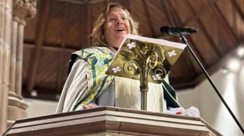 Bishop Tricia in the pulpit in the cathedral in Peel. She is wearing ceremonial robes and there are sandstone pillars to the left.