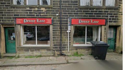 A shop bearing the words 'Penny Lane' in an old-fashioned font. A green door is to the left of the main windows, above which the name sits. A black wheelie bin is on the narrow pavement outside.
