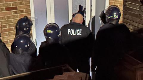 Kent Police officers wearing riot uniforms, helmets and vests stand outside a home with a pane of broken glass from the front door visible following a forced entry into the property.