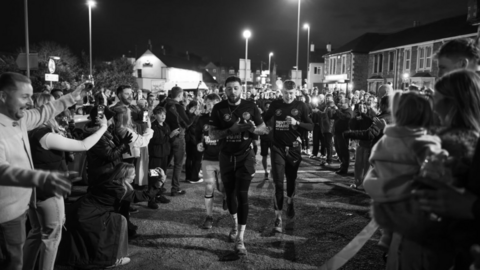 A black and white photo at night of the friends running along a residential road towards the camera, with large crowds gathered alongside, filming on cameras