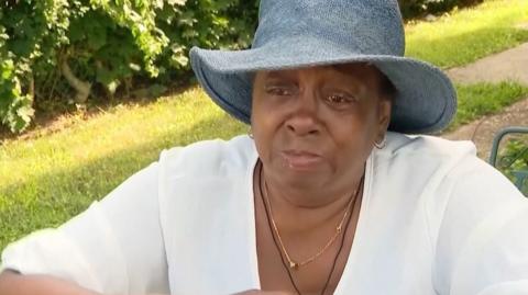 Adrienne Jones-McAllister, wearing a dark sun hat and white shirt, getting emotional in an interview.