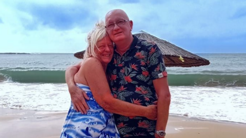An image of a blonde haired woman in a blue dress hugging a man in a floral shirt on a beach.