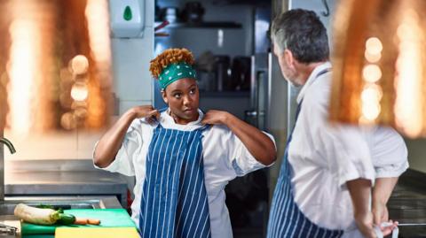 A stock photo of a trainee chef adjusting her apron in a kitchen while looking at her older mentor. There are ingredients on the counter in front of her.