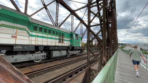 A green engine is seen on a rusty metal bridge, as a man walks alongside on a pedestrian walkway.