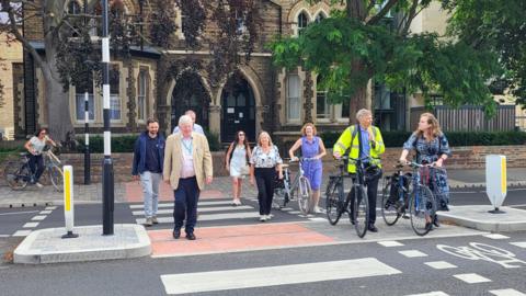 From left to right a group of people walk across a zebra crossing - Valerie Lambrechts, Joe Bates, Patrick Mulvihill, Craig Buzzard, Helen Fallon, and Councillors Judy Roberts, Louise Upton, Andrew Gant and Emma Garnett