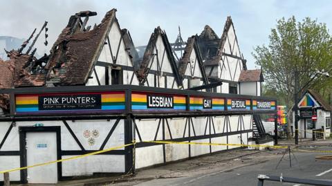 A nightclub, pictured in the middle of the day, with a roof that has been destroyed in a fire. A fire engine is parked opposite the venue. There is a long aerial ladder rising into the sky above the building. Some smoke can be seen billowing out of the venue.